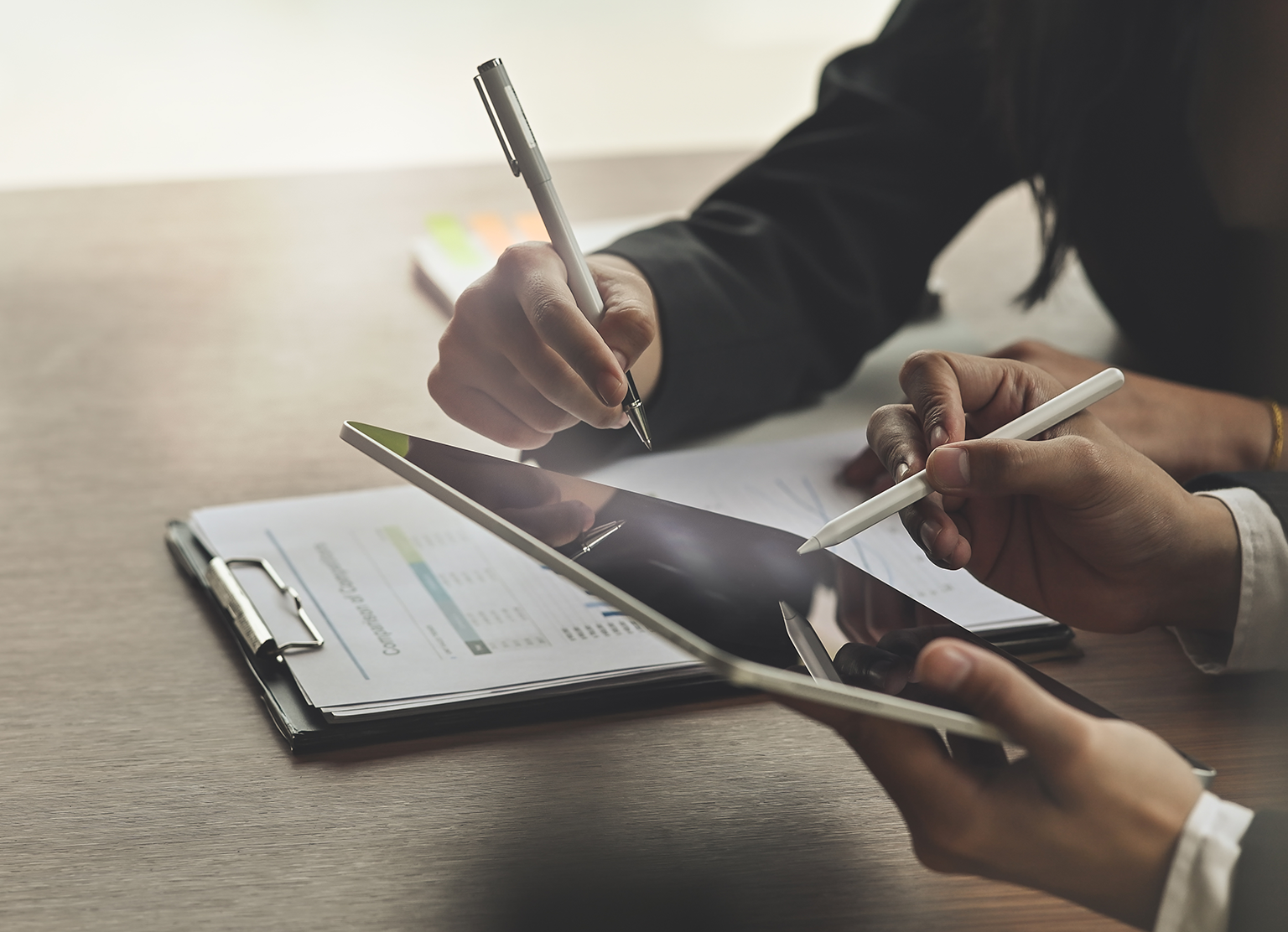 Två händer som arbetar på en surfplatta med varsin penna samt en dokumentplatta på bordet